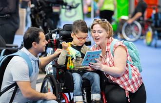 Image of a mum and dad with boy in a wheelchair, reading a book at an exhibition as a family.