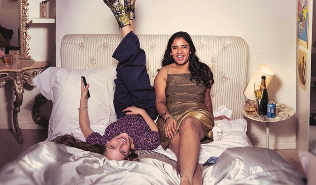 Two teenage girls sit on an rumpled bed with a pale pink satin duvet, smiling at the camera. The girl on the left lies on her back, with her feet up a
