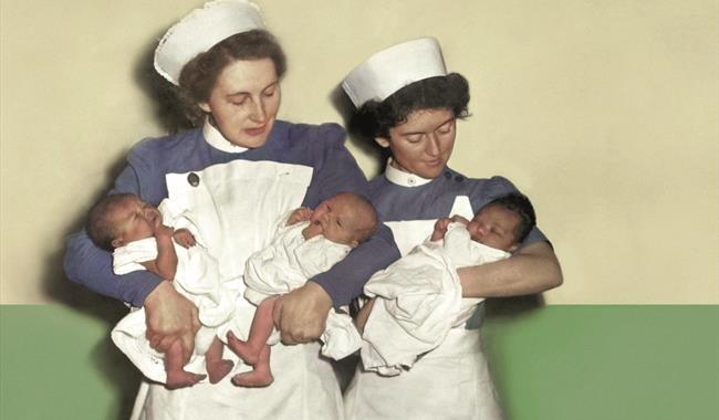 Two midwives in white smocks, white caps and blue blouses stand against a green and white wall. The midwife on the left holds two newborn babies, they