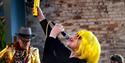 Side profile of a woman singing, looking up and holding a yellow baton, holding the microphone to her mouth. She wears a cropped bright yellow wig and