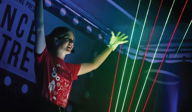 A science communicator stands with her arms in the air behind some coloured laser beams