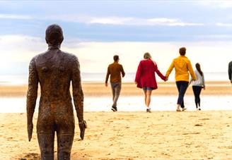 A weathered metal statue of a male is in standing in the sand looking out towards the sea. Walking towards the sea
