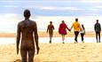 A weathered metal statue of a male is in standing in the sand looking out towards the sea. Walking towards the sea