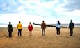 Facing the sea there is one of the weathered Gormley statues to the left of five people who are standing to mimic the statue.