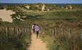 Three people are walking along a sandy walkway towards the tall, grassy sand dunes. Ahead of the dunes the sea can be seen.