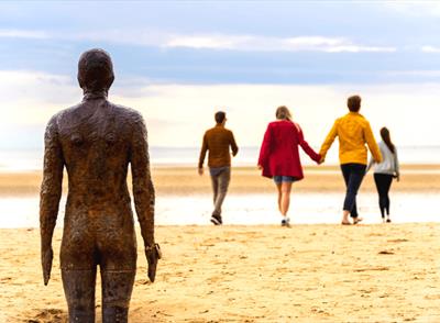 A weathered metal statue of a male is in standing in the sand looking out towards the sea. Walking towards the sea 