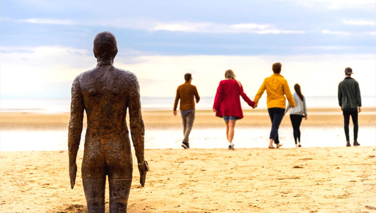 A weathered metal statue of a male is in standing in the sand looking out towards the sea. Walking towards the sea