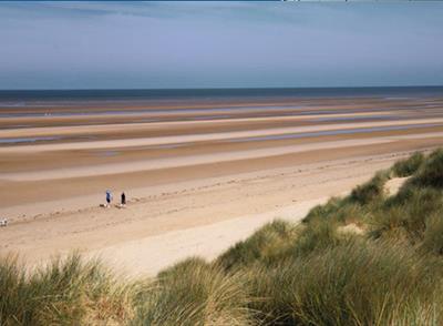 A shot of the beach taken from grassy and sandy dunes. There are two walkers on the beach. The tide is out far and blends with the blue sky.