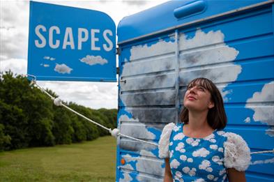 A woman in a blue dress with white clouds on it looks up to the sky, in front of what looks like a truck or van with blue skies and clouds painted on 