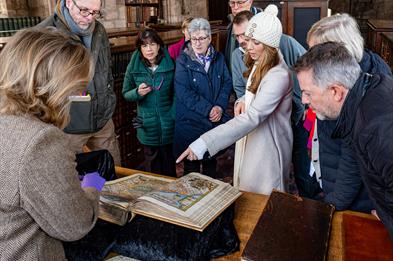 Image shows a crowd of people around an old book, with someone pointing at one of the pages