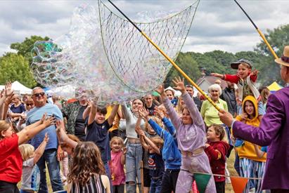 A man with a huge net creates hundreds of bubbles in front of a crowd of people, mostly children