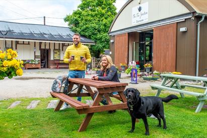 A male and female sitting at a wooden picnic bench with a dog, outside the Tea Junction Café and Manifold Information Point