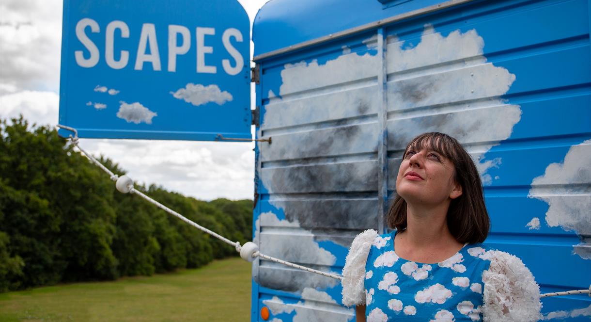 A woman in a blue dress with white clouds on it looks up to the sky, in front of what looks like a truck or van with blue skies and clouds painted on