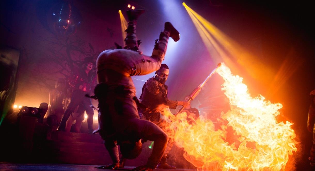 Image shows circus performers on stage, with one doing a somersault and another with a flame-thrower