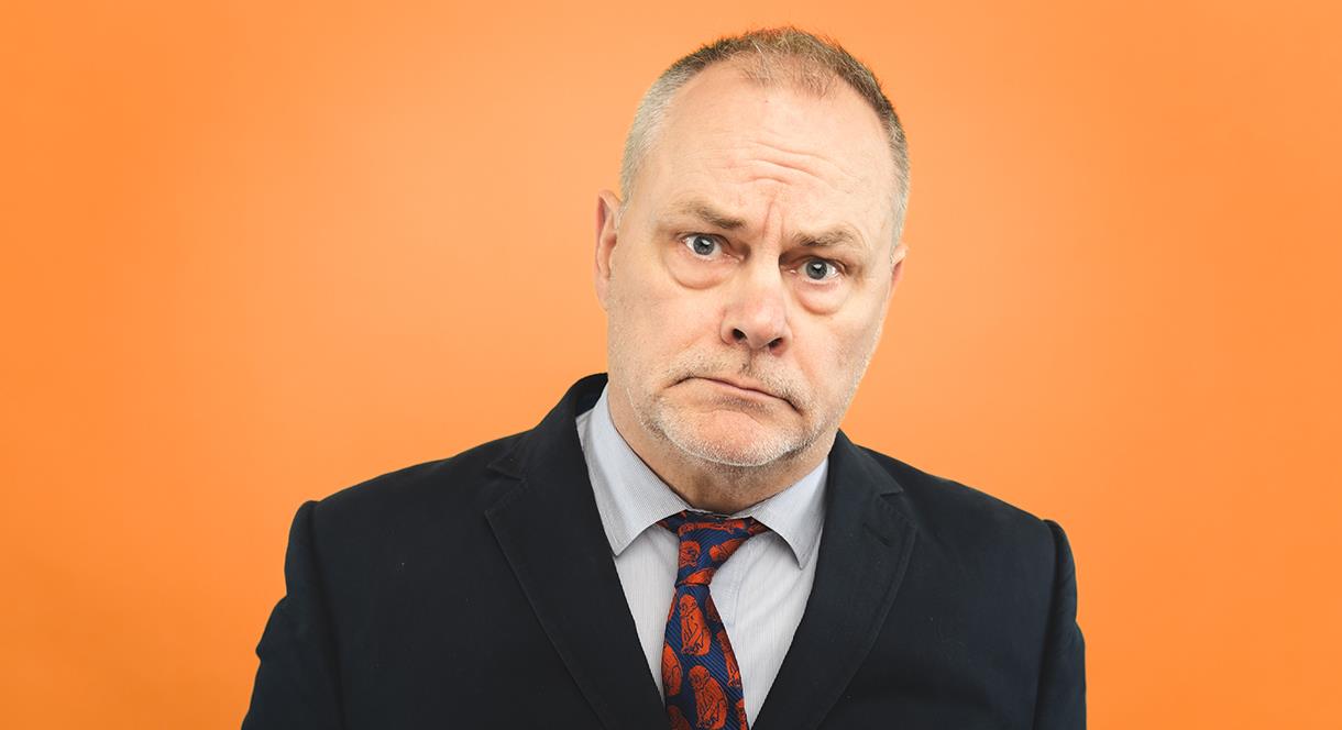 Jack Dee stands in front of an orange backdrop with his trademark miserable expression