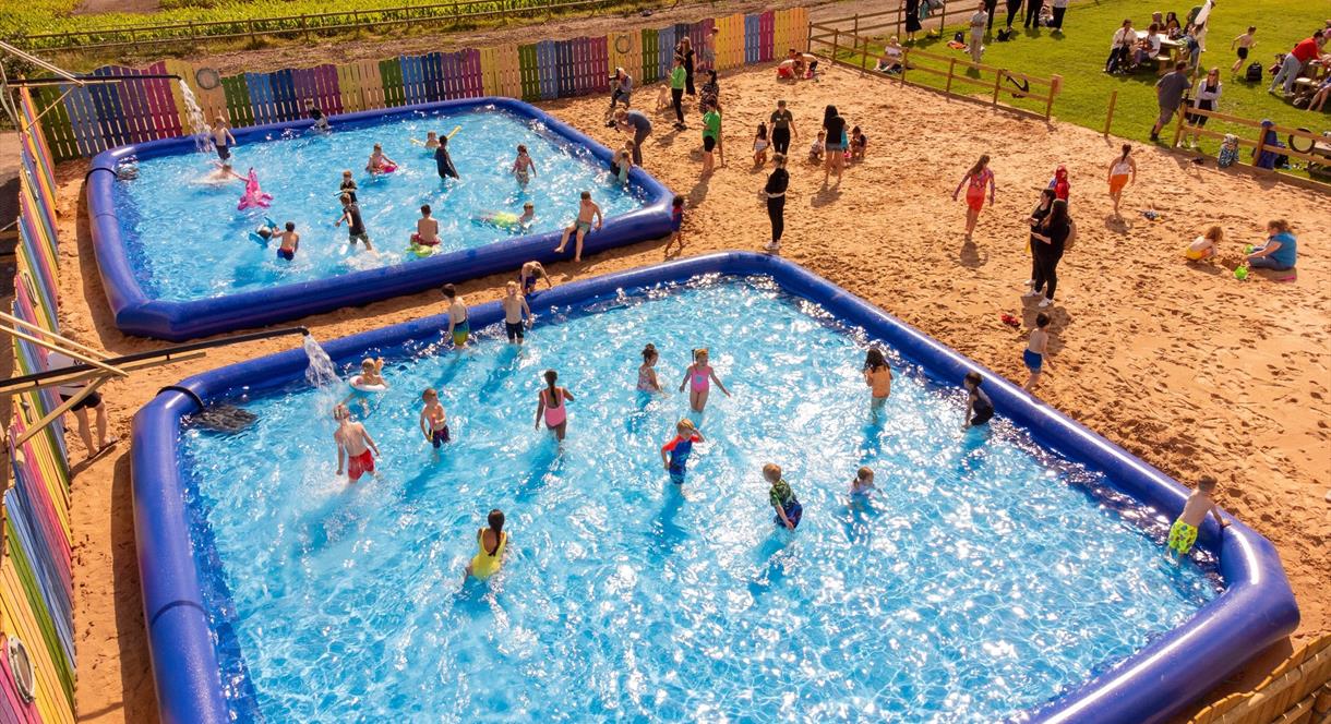 An overhead shot of children playing in two huge paddling pools, and others on a beach