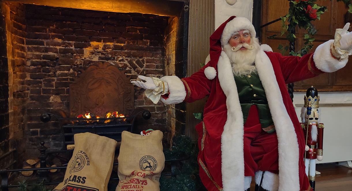 Santa sits, arms outstretched, beside a fireplace and some sacks of presents