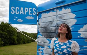 A woman in a blue dress with white clouds on it looks up to the sky, in front of what looks like a truck or van with blue skies and clouds painted on