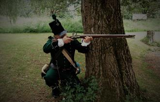 A costumed performer, as a rifleman, crouches behind a tree and takes aim
