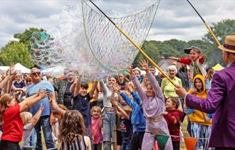 A man with a huge net creates hundreds of bubbles in front of a crowd of people, mostly children