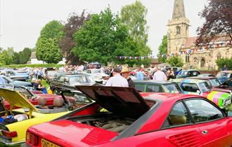 Classic cars lined up on a village green with an old church in the background
