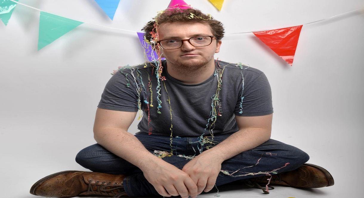 Image of a man sitting on the floor, covered in streamers and wearing a party hat