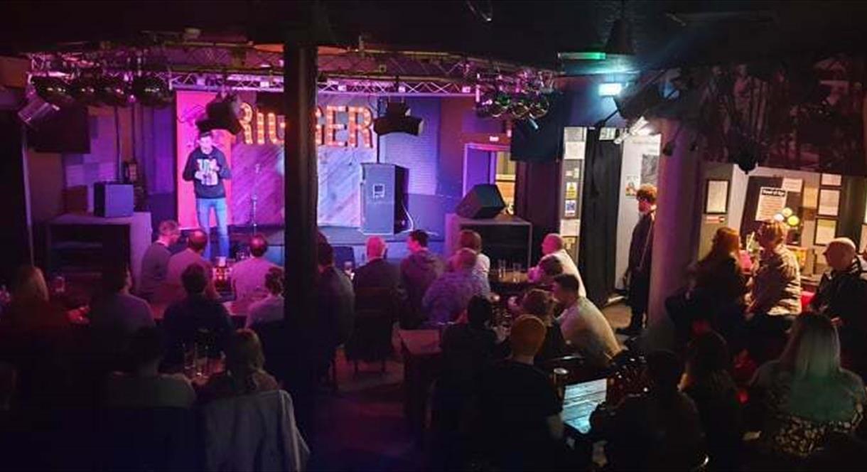 Image shows a comedian on stage at The Rigger, in front of a packed audience