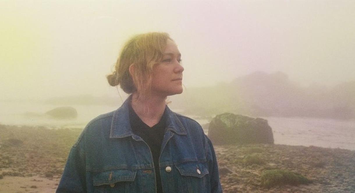 Image shows a woman, standing by a river on a foggy day, gazing into the distance