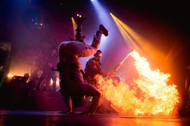 Image shows circus performers on stage, with one doing a somersault and another with a flame-thrower