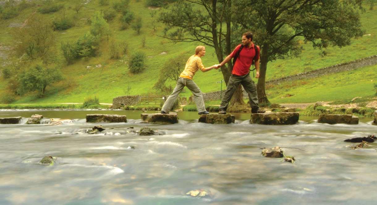 Couple crossing the famous Stepping Stones across the River Dove, Dovedale, Peak District. Staffordshire / Derbyshire border. Image courtesy Tony Plea