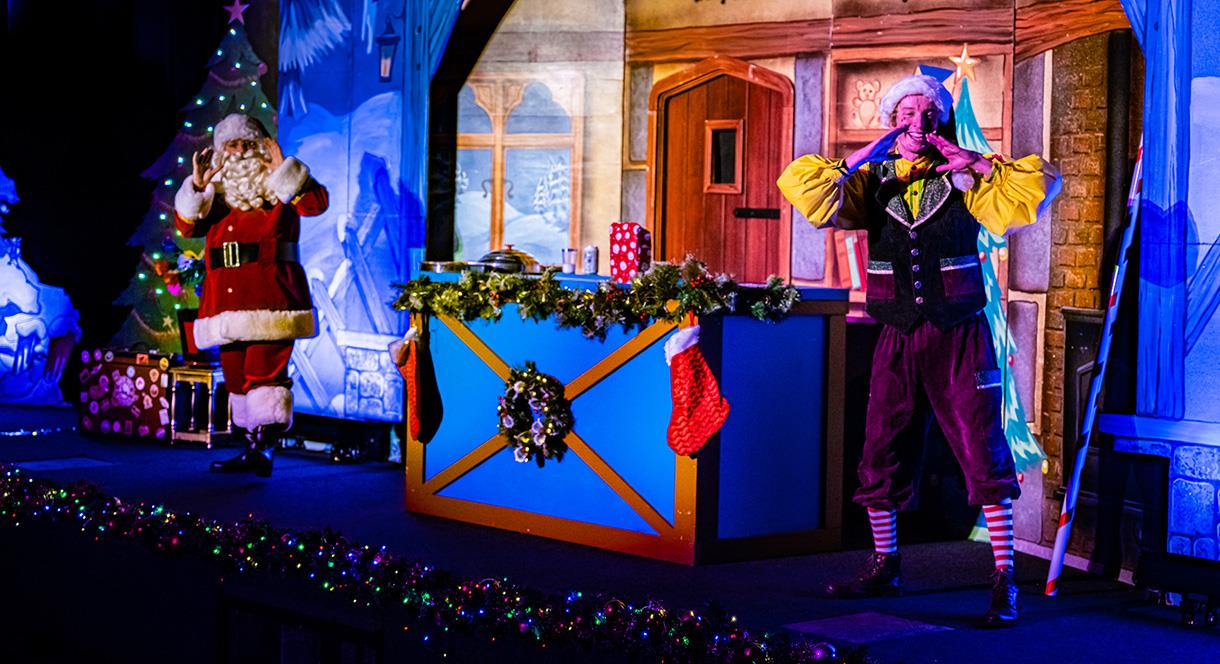 Image shows Santa and an elf performing on stage in front of a festive backdrop