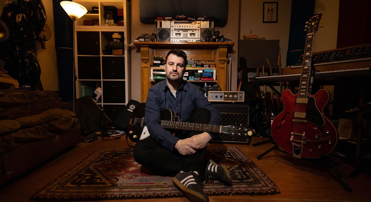 Image shows Jim Moray sitting down in his lounge with his guitars and other musical paraphernalia