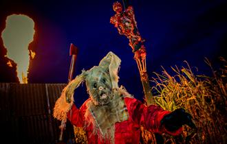 Image shows a frightening character from Screamfest, with fire shooting up into the night sky in the background