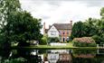 Image shows the team at the Moat House preparing the banks of the lake for an outdoor wedding. Seen from across the lake, with pretty reflections of t