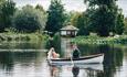 Image shows a couple taking a romantic boat trip, in their wedding outfits, across the lake. There's greenery all around the lake, and it's a cloudy d
