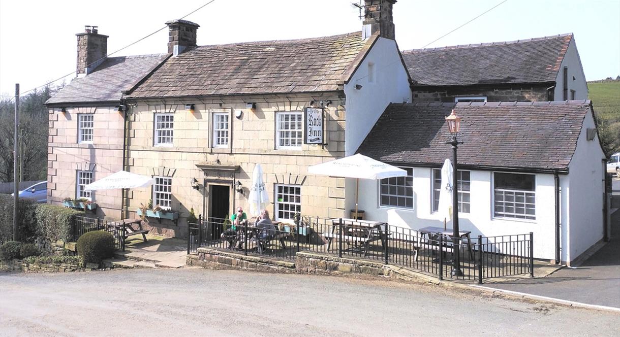 Image shows the exterior of Ye Olde Rock Inn, on a sunny day, with a few customers sitting at the tables outside