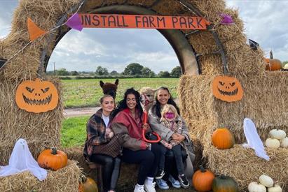Image shows a family sitting on hay bales at Tunstall Farm Park, surrounded by pumpkins, ghosts and alpacas