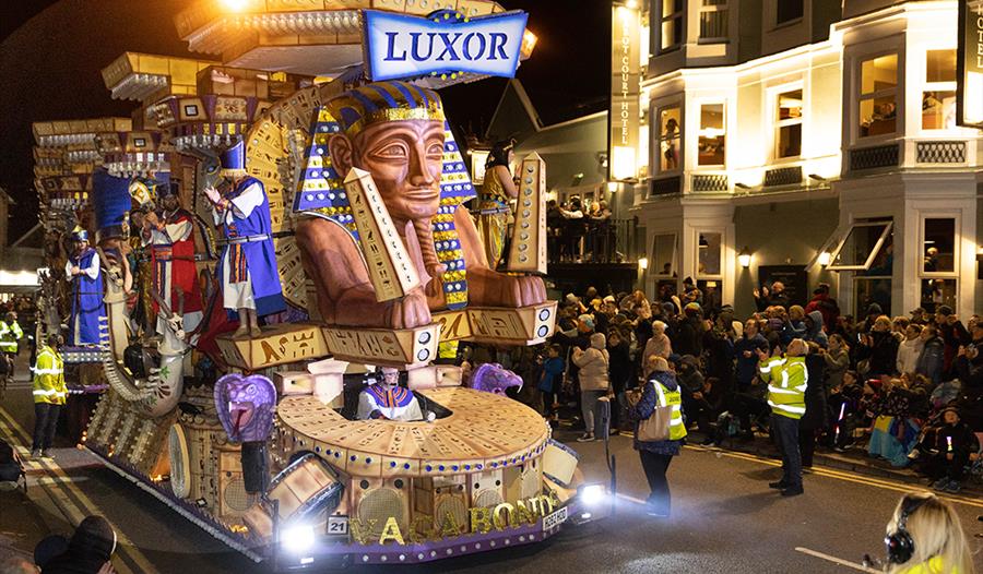 A massive Egyptian-themed huge brightly-coloured carnival float which is as tall as the buildings it is passing.