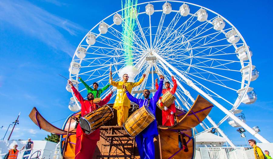 5 men playing instruments whilst dressed in colourful clothing in front of Bournemouths big wheel