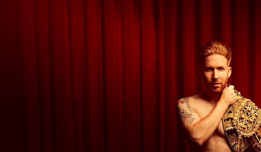 A topless man, with a Spanish matador's jacket draped over his shoulder, looks at the camera as he stands in front of a red curtain