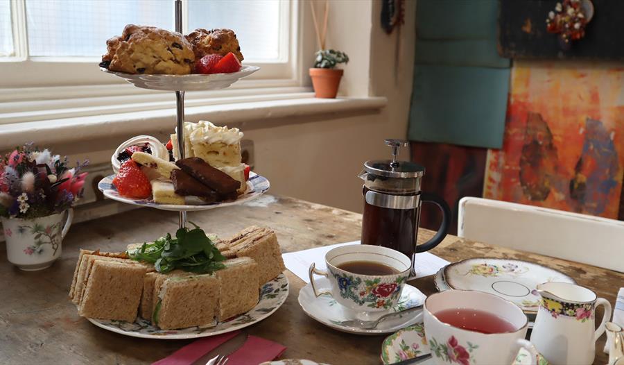 A serving of Afternoon Tea for two. A three tier cake stand showcasing sandwich squares, a mixture of cakes and fruit scones. Two teacups and saucers