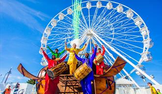 5 men playing instruments whilst dressed in colourful clothing in front of Bournemouths big wheel
