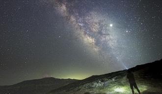 The image shows the silhouette of a man photographing The Milky Way galaxy from the hillside