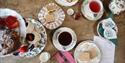 A serving of Afternoon Tea for two. A three tier cake stand showcasing sandwich squares, a mixture of cakes and fruit scones. Two teacups and saucers