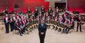 A band leader stands facing the camera, with a huge band behind him in rows, holding their instruments