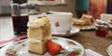 A mini two-tier cake slice with cream is in the forefront. A china tea cup and cafetière with coffee is in the background.