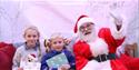 Santa sat with a young boy and a girl, both with presents. Santa is waving at the camera.