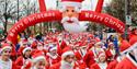 a group of runners dressed in red and blue santa suits running under an inflatable merry christmas sign.