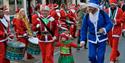 Two adults dressed as santa in red and blue suits holding the hands of a little girl dressed in a festive outfit.
