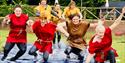 Seven people wearing togas sliding down a plastic mat covered in water.